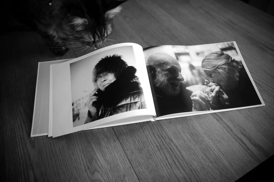 Fotobok från Crimson placerad uppslagen på ett träbord. I uppslaget finns två svartvita bilder med porträtt av människor. Bredvid fotoboken siter en katt.