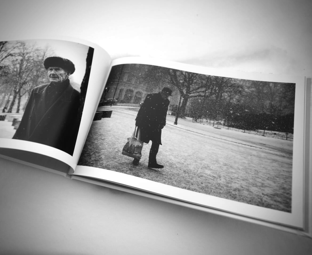 Fotobok från Crimson lagd uppslagen i snö. Fotoboken innehåller svartvita fotografier av en man som promenerar i stadsmiljö.
