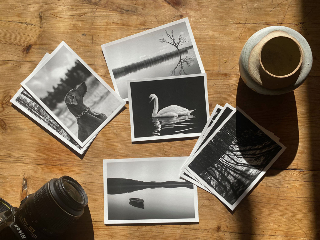 Utskrivna svartvita bilder med vit kant som ligger på ett träbord. I bildkant finns en kamera och en tom vas.