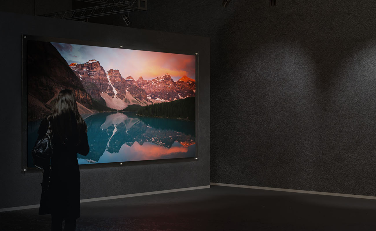 Ett stort direktprint på plexiglas med motiv av berg i rött solljus sitter uppsatt på svart gallerivägg. En person står och tittat på bilden.