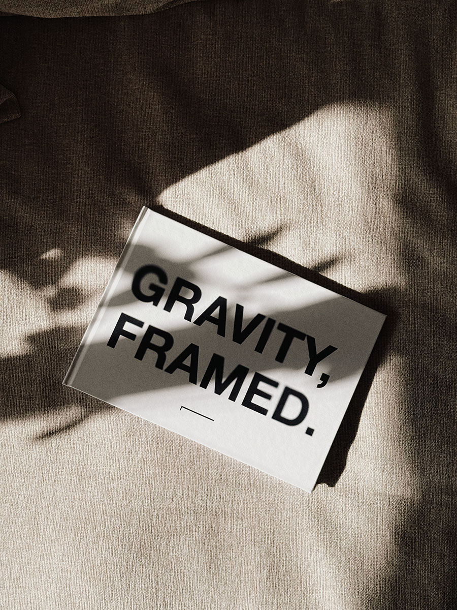 En vit bok som med texten Gravity Framed, liggandes på en beige soffa med skuggor över bilden.